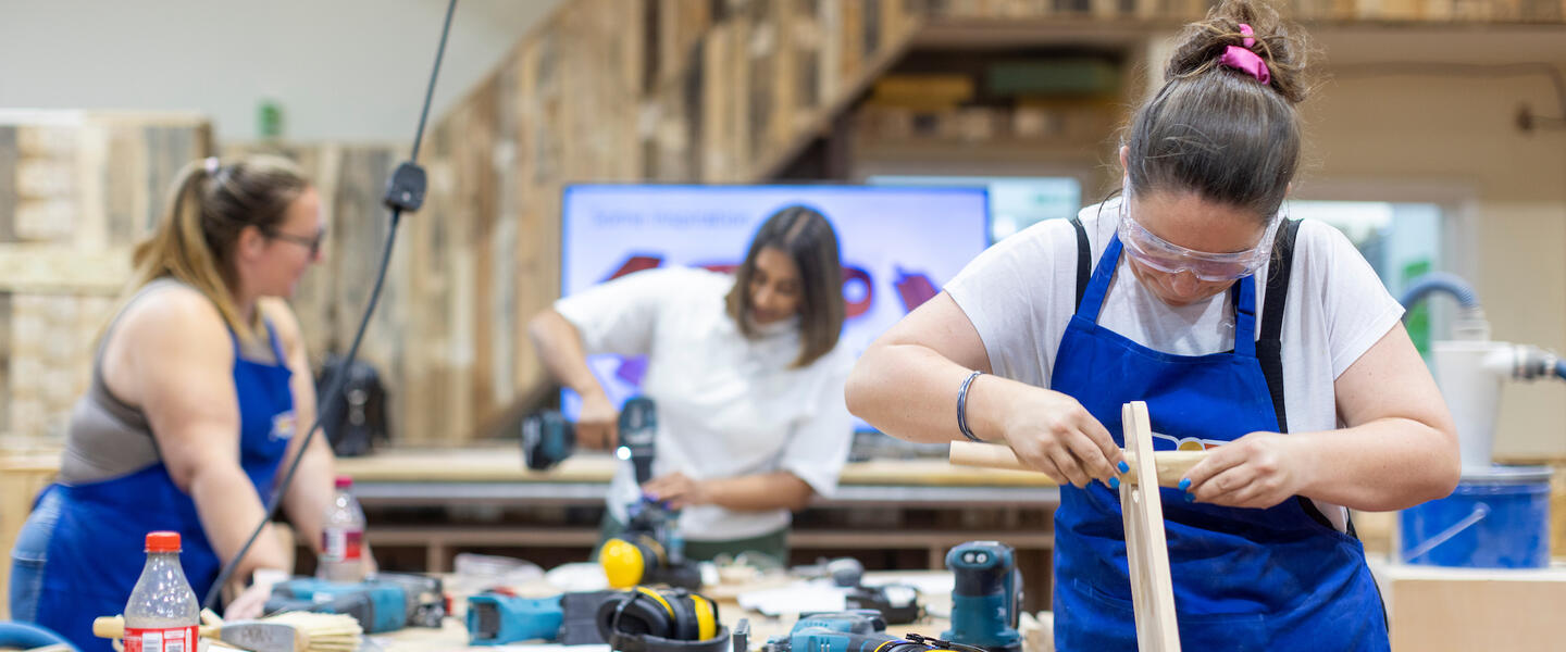Three women working in a workshop with power tools and timber